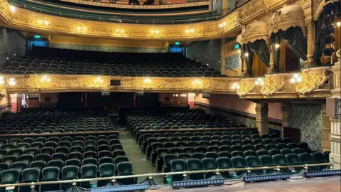 The view from the stage of the theatre, you can see a small section of the brown stage. Rows of green velvet seats make up the stalls, and more can be seen in the circle too. There are individual boxes close to the stage on the circle-level. Ornate designs can be seen on the outside of them in a cream colour, which connect to front of the balconies.