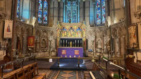 Image of the alter at the front of a church. The alter is dressed in purple and the walls are adorned with gold artwork.