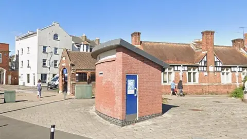 The picture shows a small brick building with a curved roof and a blue door sitting in an open paved area. It looks like an old public toilet block. Behind it there are older red‑brick buildings with tall chimneys and black‑and‑white timber detailing, along with a larger pale-coloured building on the left. A few people are walking across the open space, and the scene is bright with clear blue sky.