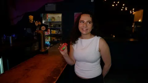 Jessica Unger is holding a Lift glucose shot next to a bar at a club. She wearing a white vest and smiling. The room is quite dark. The beer drafts, pantry and fairy lights in the background brighten up the room a little.