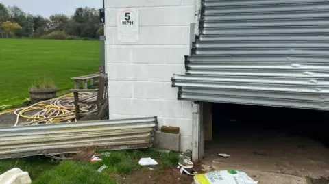 Broken shutter doors on an outbuilding are lifted or pulled out halfway with debris on the floor outside next to some cables and rubbish.