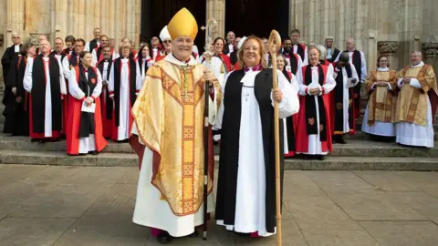 Duncan Lomax The Reverend Patricia Hillas stands next to Stephen Cottrell, Archbishop of York, outside York Church. They are both dressed in priestly garb and holding a staff. Other members of the clergy line up on the steps behind them.