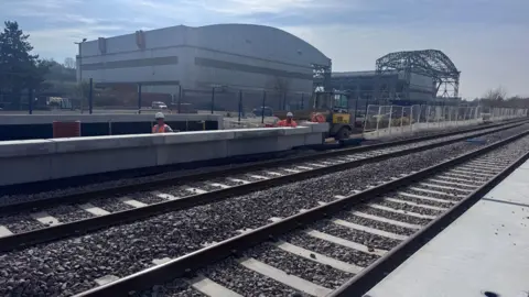 Some railway lines in the foreground and the Bristol arena buildings behind. The picture is taken on a sunny day and the arena buildings are large and grey