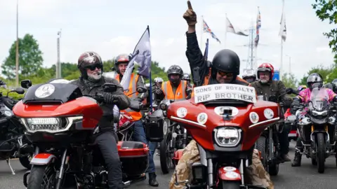 Hairy Biker Si King on his motorbike at the National Motorcycle Museum in Solihull during Dave Day. Next to him is another rider who is raising his arm and pointing his index finger in the air. Behind them are other bikers.