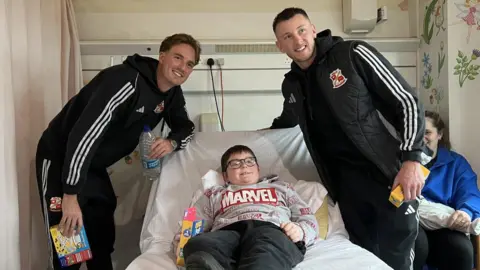 two Swindon Town footballers leaning over a hospital bed with a child in it - child wearing a comic book hero jumper - woman slightly hidden in background 