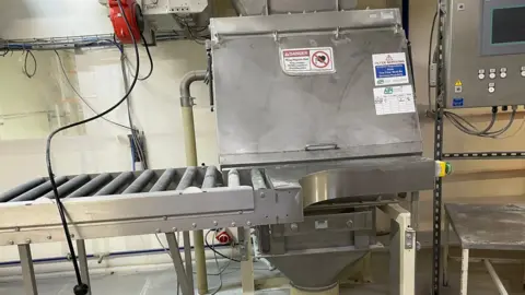 HSE A close up of an industrial food-production area containing stainless-steel processing equipment and a roller conveyor system. Above and beside the equipment are electrical cables, pipes and ducting. 
