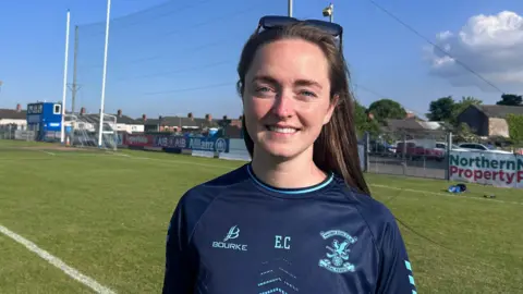 Eimhear Costello, standing on a grass pitch, she has long brown hair and is wearing sunglasses on her head. She has a dark blue and light blue trimmed short-sleeved jersey/t-shirt on with Bourke written on one side, E.C in the middle and the clubs crest on the other side. 