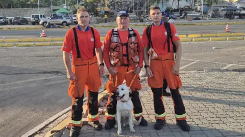 ECFRS Rob Wingar, Graham Currie and Richard Swash all wearing orange tops and trousers. They are standing in front of a road and there is a collapsed building behind them. Bailey, a white labrador, is sitting in front of them.