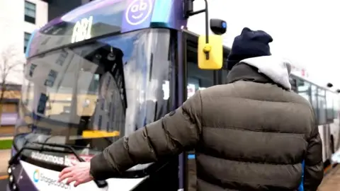 PA Media A man wearing a black padded jacket and a navy hat holds out his arm to hail an autonomous bus marked AB1. 