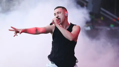 A man in a black vest holds a microphone to his mouth in his left hand while pointing out with his right hand, his arm outstretched. He has close-cropped dark hair.