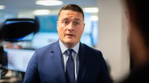 Wes Streeting speaks to a journalist at NHS England's headquarters wearing a blue suit and tie.