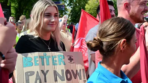 A young woman with long blonde hair holds a brown cardboard sign which says 'better pay now, fair pay to guard your day'. She is wearing a black t-shirt and a silver cross necklace. There are other men and women surrounding her holding red union flags. They are wearing red, blue, white and other coloured clothing.