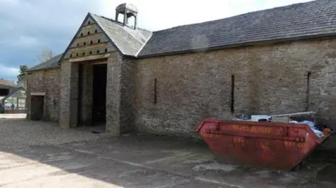 Monmouthshire council A large stone barn with a red skip full of rubble outside a wooden planked door 