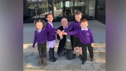 Four children in purple jumpers and a man in a black blazer all hold a key infront of a building with glass doors and windows behind. 