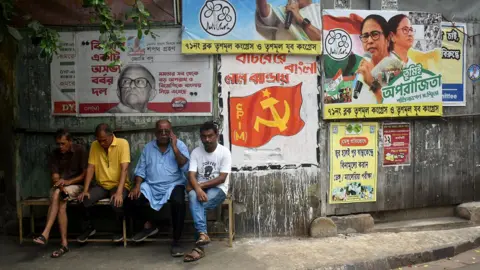 A group of people sit on a bench next to political party campaign posters on a wall on a street in Kolkata on 29 March