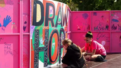Rob Harris/ Metal Peterborough Two artists sit on the ground and paint on a large piece of artwork that is hanging on a bright pink wall.