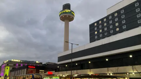 St Johns Beacon against a cloudy sky with St John centre building at ground lever lit up with red lights.