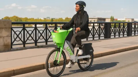 A woman dressed in a black jumper and black jeans is riding a green and white lime bike. She has braids and wears a black helmet. She is riding across a bridge with the skyline of the city and river in the distance.