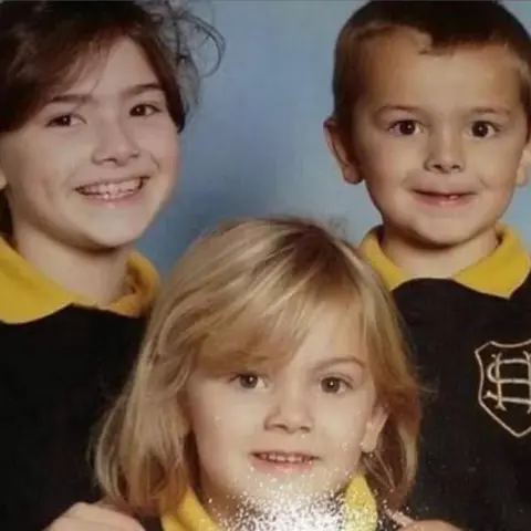 A school photo of three children in uniform. They are wearing dark jumpers over yellow polo shirts. 