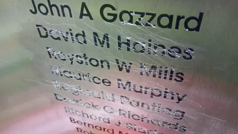 Black names engraved onto a silver metal plaque with scratches all over them, as if an attempt has been made to scratch them out.