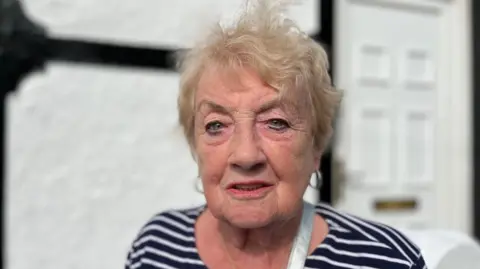 Christine looking at the camera. It is a head and shoulders shot of her. She has wispy blonde hair and green eyes. She has pink lipstick on and you can see the silver shoulder strap of a bag on her left shoulder. She has a blue and white striped top on. Behind her is a blurred image of a white door with a gold letterbox.