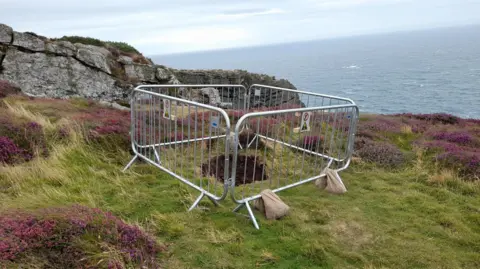 Four silver safety barriers sit around a hole in the ground in an area close to the edge of a grassy area, with patches of purple heather. 