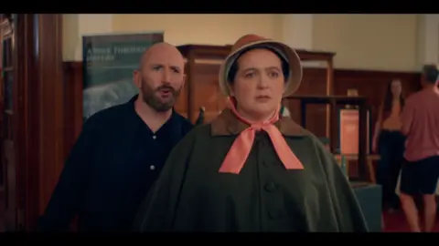 A woman in a bonnet looks away as a bald man with a beard talks behind her.