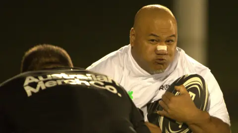A man wearing a white shirt while holding a rugby ball runs at another man wearing a black shirt who is preparing to tackle him