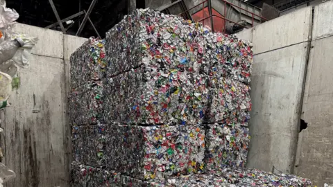 Stacks of tightly compressed bales made from crushed aluminium cans are piled high inside an industrial waste‑processing facility. The bales form large rectangular blocks against tall concrete walls, with loose debris scattered across the floor.