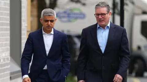 Sir Sadiq Khan and Sir Keir Starmer walking side-by-side on a public street. A lorry is seen out of focus in the background and they are next to a grey bricked building.