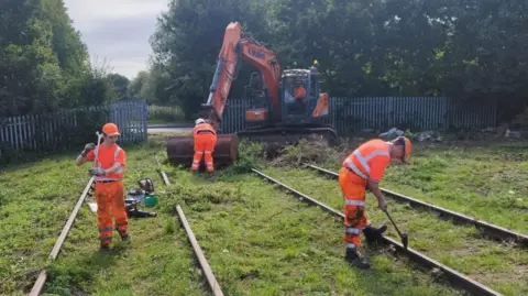 Three men in bright orange overalls lifting railway track with the help of a digger