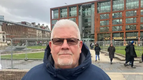 A man with short grey hair and a beard is wearing glasses and a dark blue coat while standing close to railings in Piccadilly Gardens. Around six people can be seen walking nearby.
