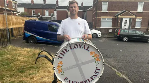 A man in a white top is looking at the camera holding a white drum saying in purple writing, "The Loyal East Belfast". He is standing in a residential street with parked cars in the background.