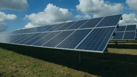 Sunnica A view of large solar panels on legs set into close-cropped grass with a blue sky with white clouds above