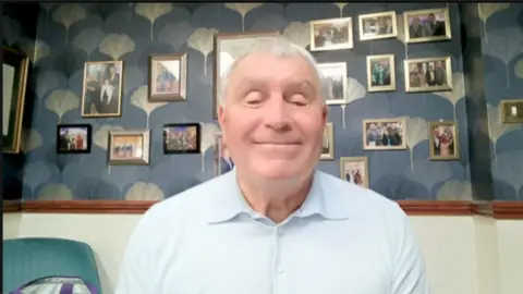 Peter Shilton in a room with lots of small, framed photographs behind him on the wall. He is smiling and looking straight at the camera.