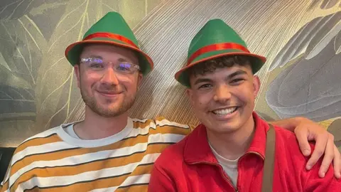 Gareth Scourfield One man in yellow and white stripy t-shirt wearing a red and green hat, with his arm around another man who has a red top on and the same red and green hat