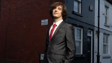 PA Media Brad Kella wears a dark suit with a red tie as he stands in a Liverpool street of terraced houses with a sign on the wall reading "All ball games prohibited"