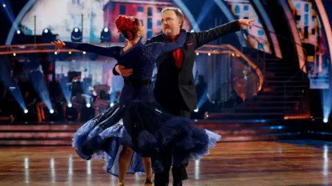 PA Media Chris McCausland, in a black blazer over a red shirt, dances with Diane Buswell. Both have their left arms outstretched behind them and hold on to each other with their right