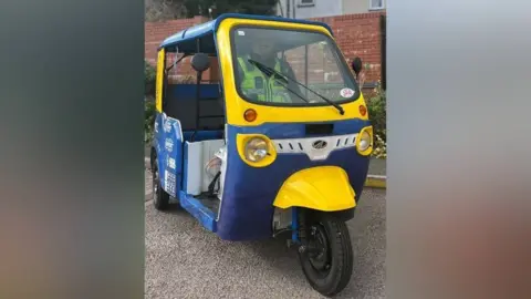 A blue and yellow painted tuk-tuk is parked on a road. A police officer is inside, wearing a high visibility jacket. Behind him are plants and a brick wall.