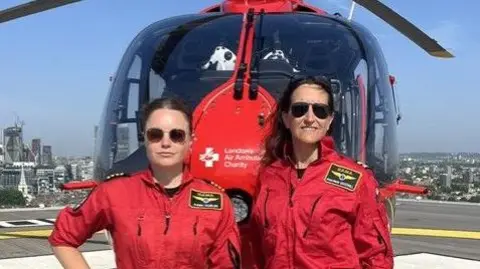 Dobler (left) and Goodwill (right) looking straight into the camera with neutral expressions, both wearing their red uniforma nd sunglasses, with the red helicoper right behind them and a London skyline in the background.