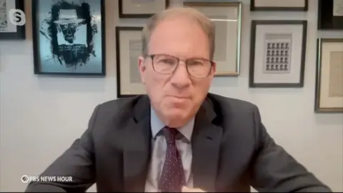 Jeffrey Goldberg is doing a virtual interview with PBS News Hour, sitting at a desk with frames behind him.