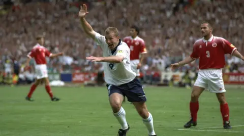 Getty Images Alan Shearer puts hits hands up in celebration after scoring a goal. Behind him are three football players looking shocked.