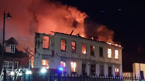 Large orange flames and smoke rise from the Angel inn which is two storeys tall and has large words reading "Angel Inn" on the front. Fallen roof timbers can also be seen and a fire engine out the front