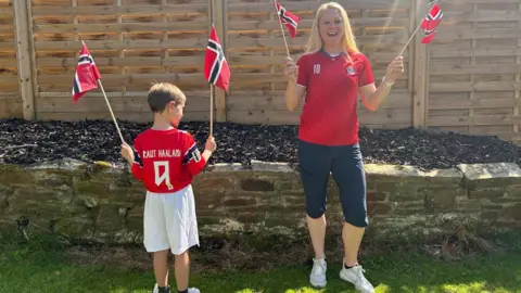 Julia Bryson/BBC A woman and her son, bith in Norway football tops, in their garden waving Norweigan flags. 