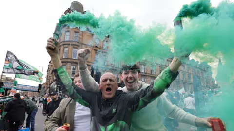 PA Media Three Celtic fans cheer for the camera while celebrating winning the league title at the weekend. One fan, a bald man in a track suit, has two smoke cannisters in his hand, which are letting off green smoke.