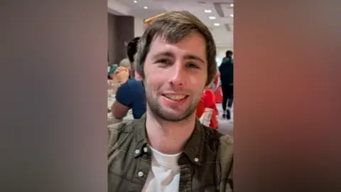 Carl, who has brown hair with a medium length fringe, smiles at the camera. He is wearing a greeen shurt and white t-shirt.