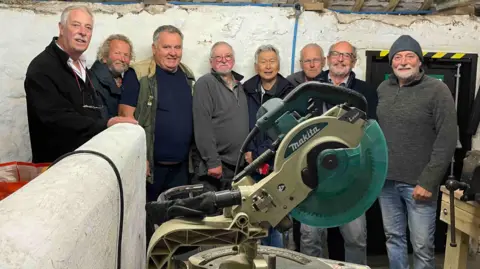 Eight men at the Men's Shed St Just in Penwith. They are standing in a line behind a green Makita table saw.