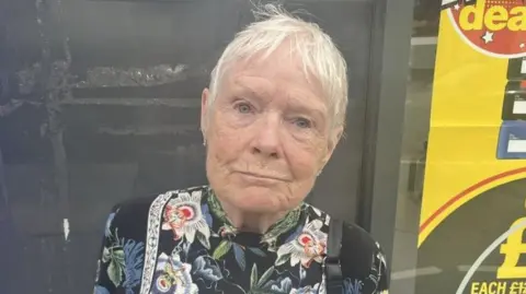 BBC/Rachel Russell A woman with short white hair looks into the camera. She is wearing a black top with a floral pattern and is standing outside a shop.
