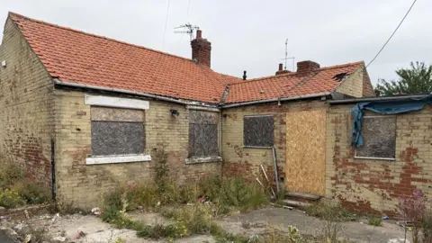 The back of the derelict bungalow has all the windows boarded up. Some boarding looks newer than others. Long grass has grown up through cracks in the paving outside.
