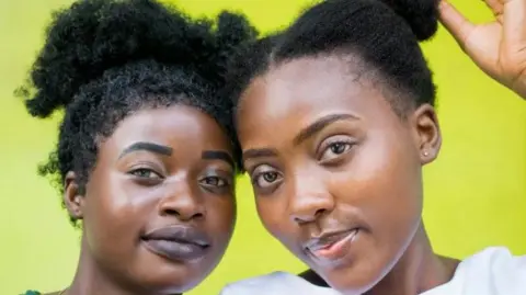 A head-and-shoulders close-up of two women with black afro hair posing with their heads close together against a bright yellow background. One has her hand to her hair.